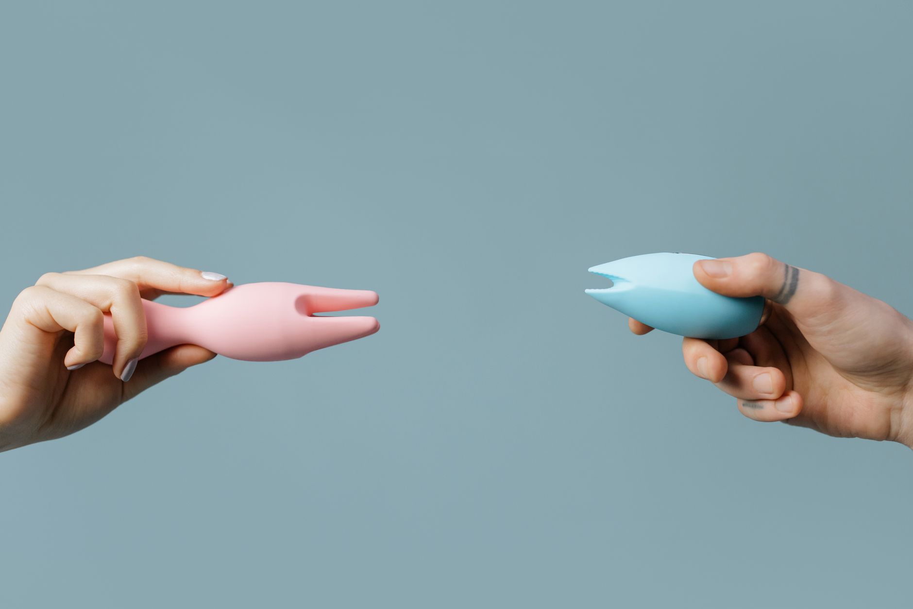 Two hands holding pink and blue silicone vibrators together, representing partnership and shared pleasure.